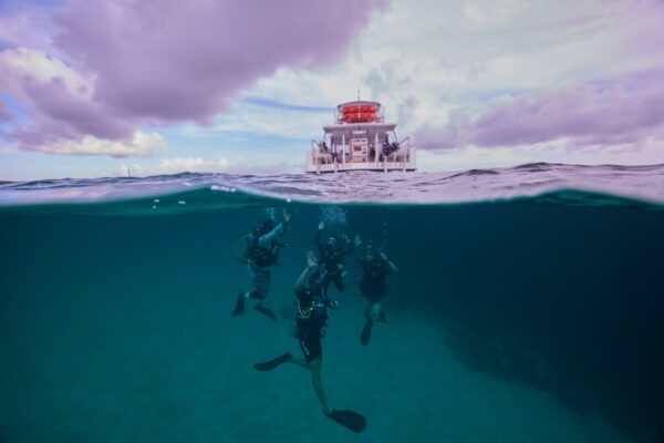A scuba diving instructor and her students surface near a boat with one arm raised each to check for items on the surface.