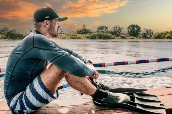 a diver sits on the seashore gazing out over the ocean. He is wearing his fins and simming trunks and a baseball cap.