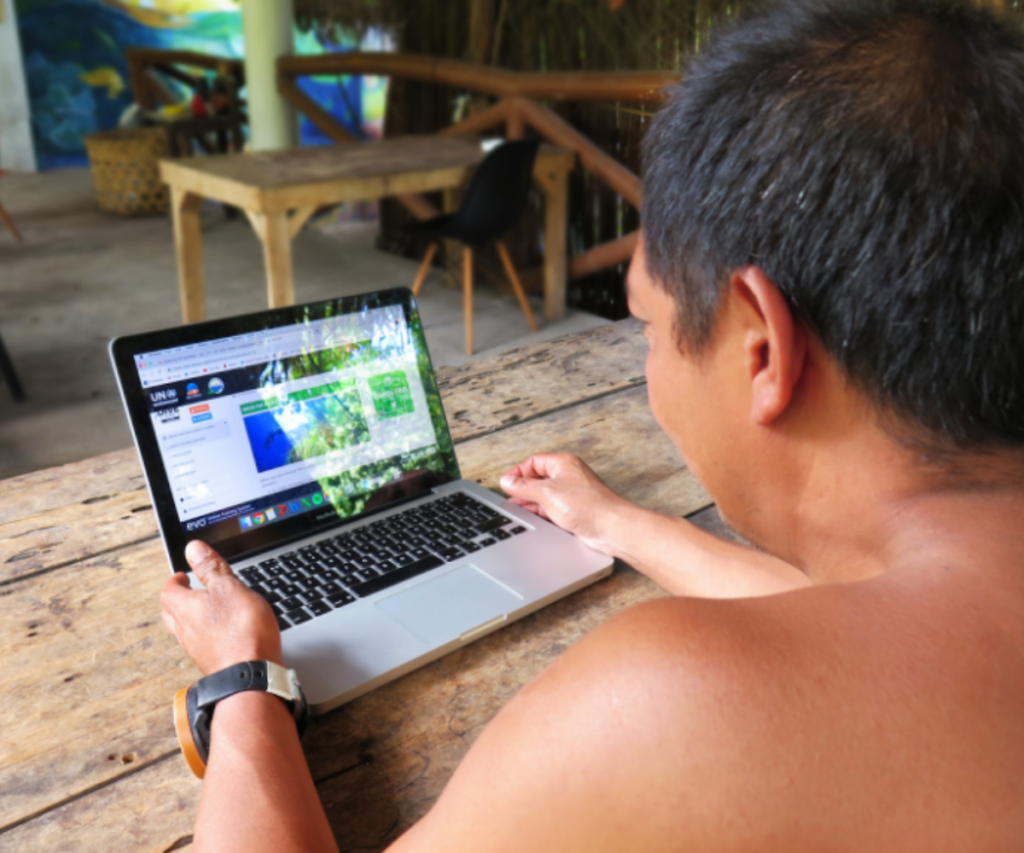 a man is sitting in a dive shop looking at the Green Fins Community hub on his laptop