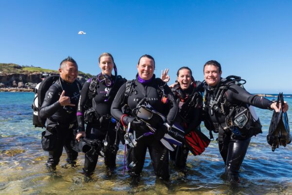 A group of divers poses for the camera while exiting the water onto the beach