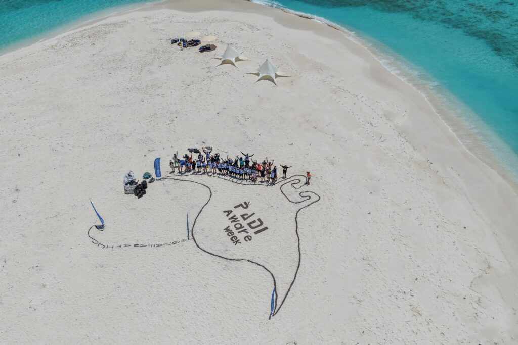 Аэрофотоснимок дайверов, празднующих PADI AWARE Week 2025 на Мальдивах, на фоне нарисованного на песке ската манты