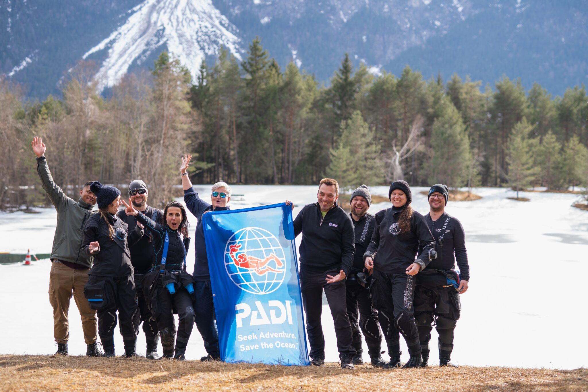 A group of people stand on the ice on a lake in Austria after going ice diving. They are holding a PADI flag.