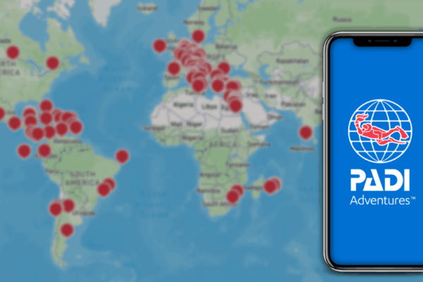 Smartphone displaying the PADI Adventures app in front of a world map marked with global scuba diving locations, highlighting international dive sites and bookable adventures.